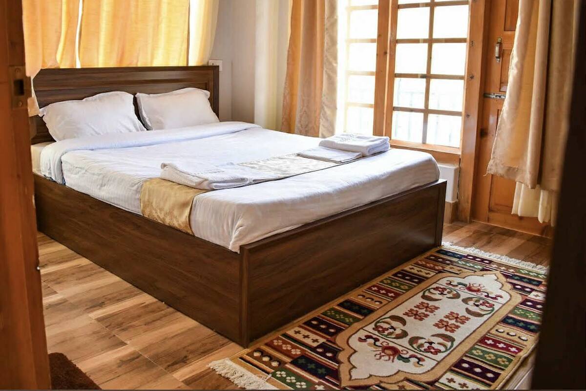 Una cama de madera con ropa de cama blanca, una alfombra estampada y una ventana con cortinas.