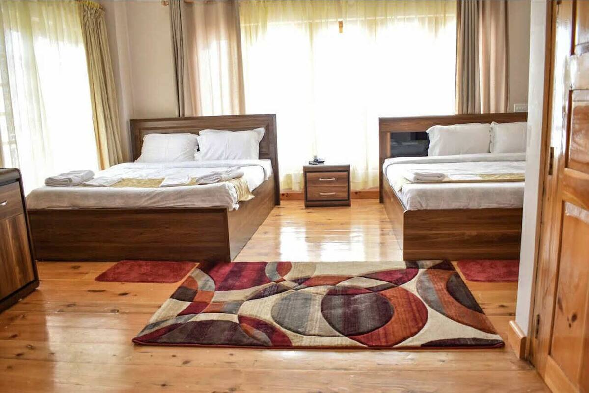 Habitación de hotel con dos camas, cabeceros de madera, mesitas de noche pequeñas con lámparas y una alfombra abstracta grande sobre el suelo de madera.