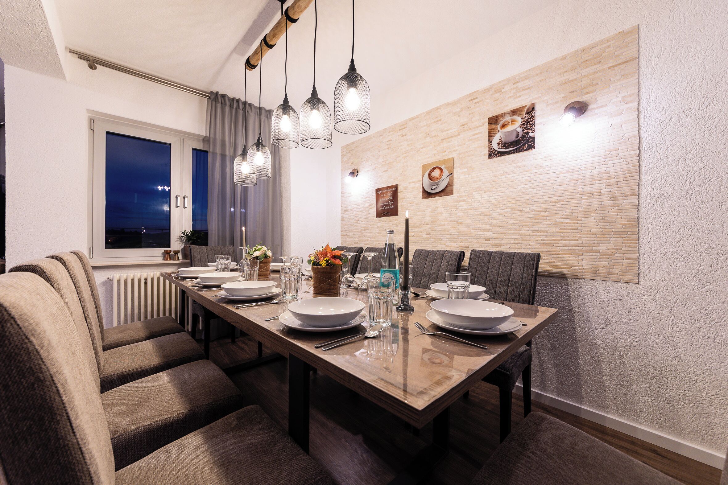 A dining area with a long table set for four, a window with curtains, and a wall-mounted light fixture.