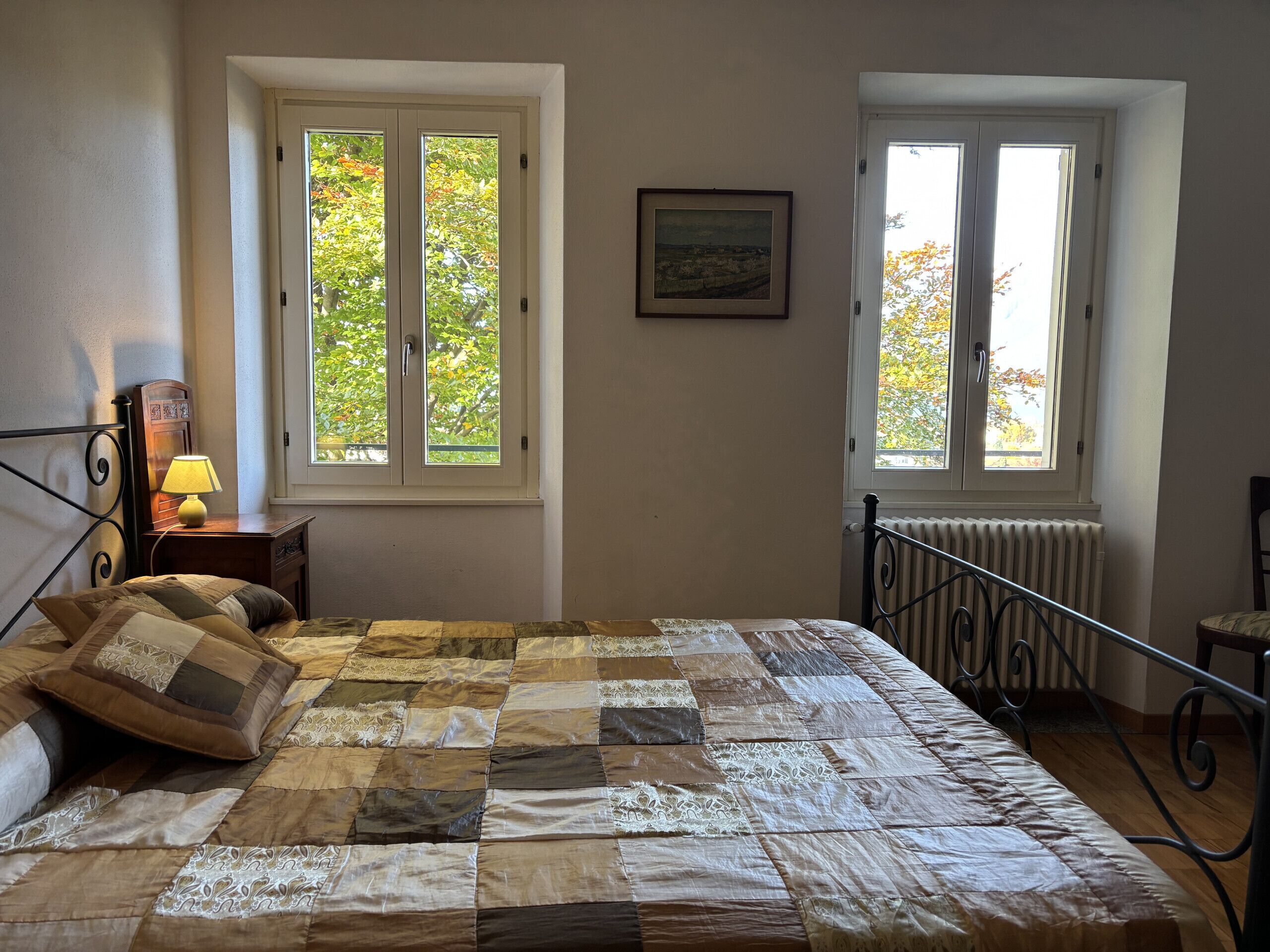 A bedroom with a large bed, two windows, a bedside table with a lamp, a framed picture on the wall, and a radiator.
