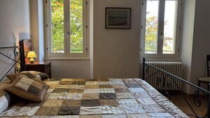 A bedroom with a large bed, two windows, a bedside table with a lamp, a framed picture on the wall, and a radiator. - Esther 2 apartment with balcony in Luino (Luino)