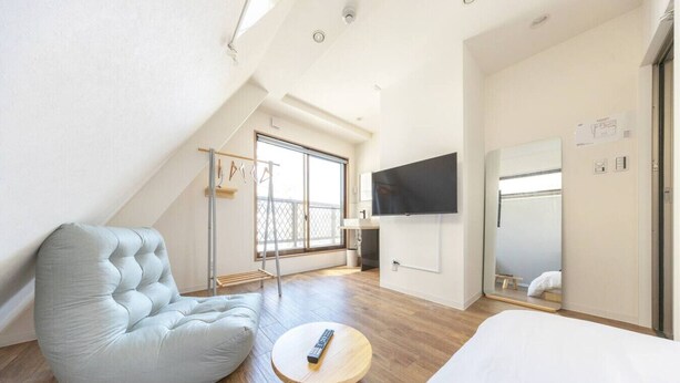 A modern room with a sloped ceiling, a large bed, a seating area with a chair, a small round table, a TV mounted on the wall, and a balcony with a railing. - The best Nakano house (Nakano City)