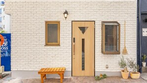 A building with a white brick facade, a wooden door with a triangular window, a bench, and two potted plants. - The best Nakano house (Nakano City)