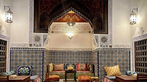 Un intérieur marocain traditionnel, caractérisé par des motifs complexes de carreaux, un espace de détente central agrémenté de coussins et des meubles en bois.