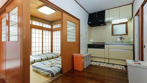 A compact room with a futon bed, a small kitchen with a dishwasher, and a sliding door leading to another room. - House Near Shinjuku Central Park! (Shinjuku-ku)