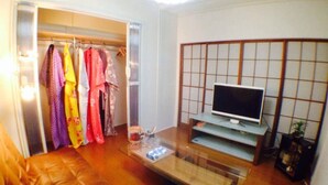 A compact living space with a glass coffee table, a TV on a stand, a wardrobe with colorful clothes, and a traditional Japanese shoji screen. - House Near Shinjuku Central Park! (Shinjuku-ku)