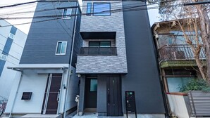 Un edificio residencial moderno de dos pisos, con fachada gris oscura, un pequeño balcón en el nivel superior y una puerta principal negra.