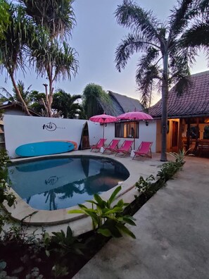 A swimming pool with lounge chairs and umbrellas, surrounded by greenery and a building with a tiled roof. - Ardi Beltza Villa (Gili Air)