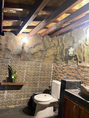 A bathroom with a rustic stone wall, a white toilet, a wooden shelf with a vase of flowers, and a stone sink. - Ardi Beltza Villa (Gili Air)