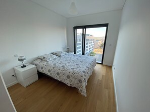 Une chambre à coucher avec un lit, une table de chevet, une lampe et une vue sur les immeubles par une grande fenêtre.