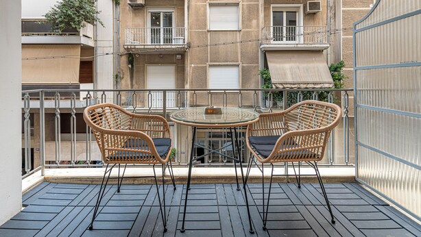 A balcony with two wicker chairs and a glass table, overlooking a building with multiple balconies and windows. - Alopece City Apartment (Athens)