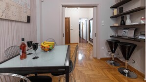 A dining area with a glass table, chairs, a bottle, glasses, and a fruit bowl. There are two black bar stools and a shelf with decorative items. - Alopece City Apartment (Athens)