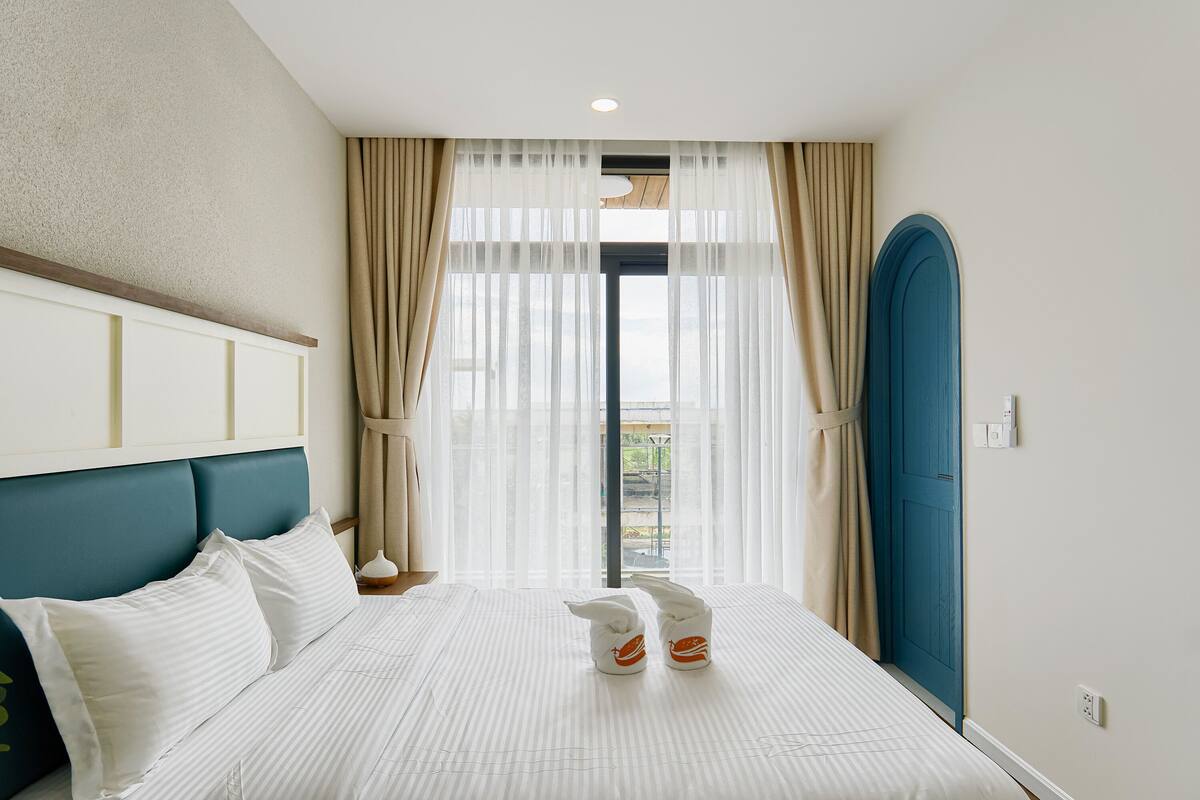 A hotel room with a bed, curtains, a blue headboard, and a view through the window.