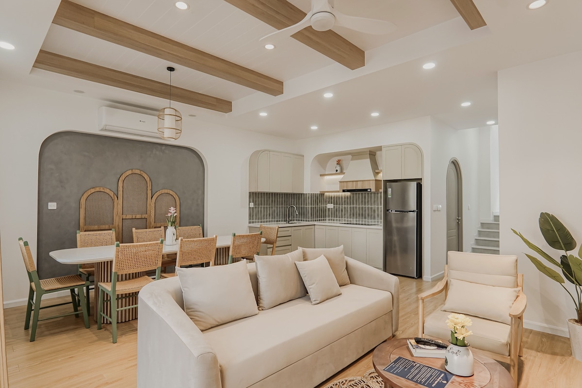 A modern living room with a sofa, dining table, chairs, and a kitchen area with a refrigerator.