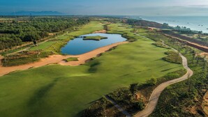 An aerial view of a golf course with a large pond, surrounded by green fairways and sand bunkers, with a coastal view in the background. - Life Travel Villa - Novaworld Phan Thiet (Tien Thanh)