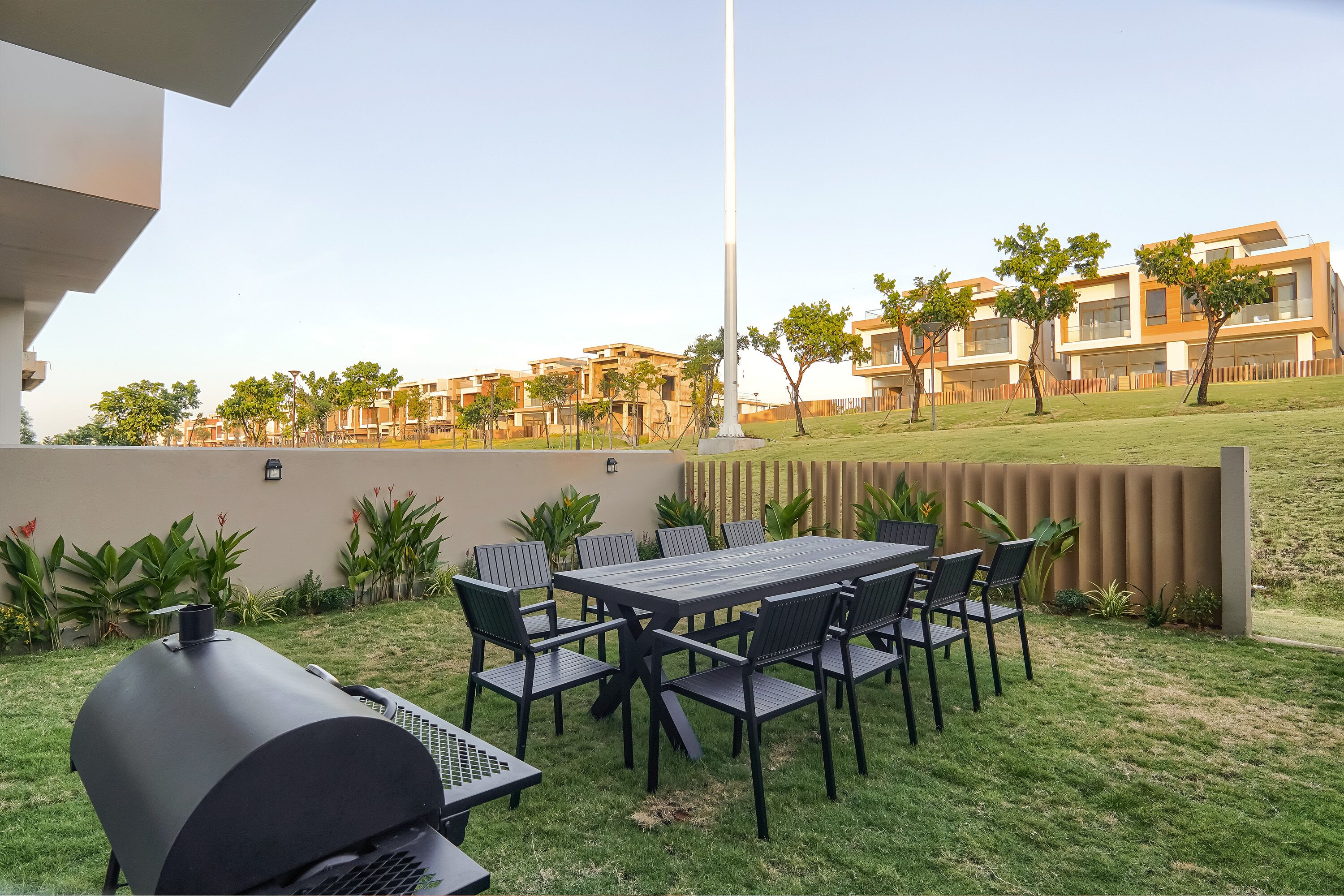 A patio with a grill, table, and chairs, surrounded by a fence and greenery, with modern buildings in the background.