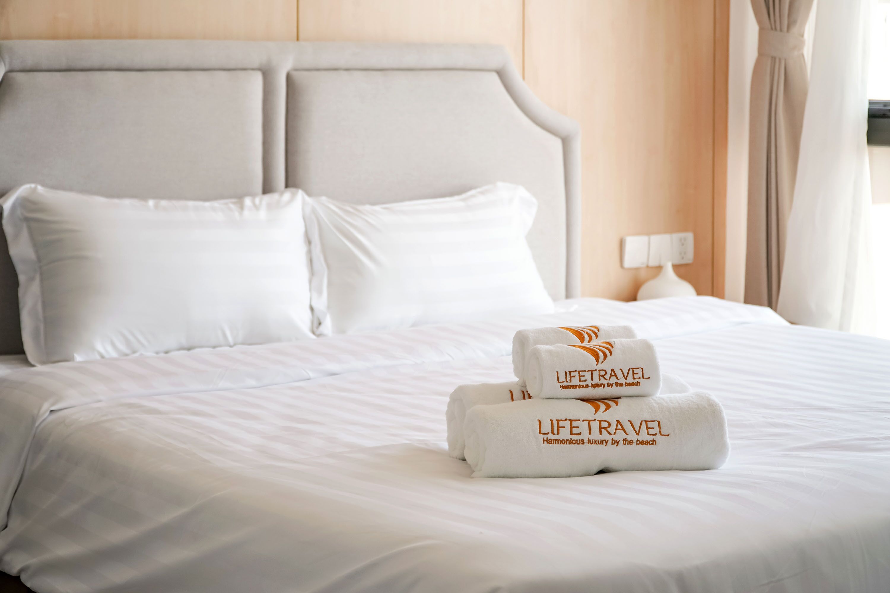 En snyggt bäddad säng med vita lakan, två kuddar och två handdukar med logotypen ”LIFETRAVEL” i ett hotellrum.