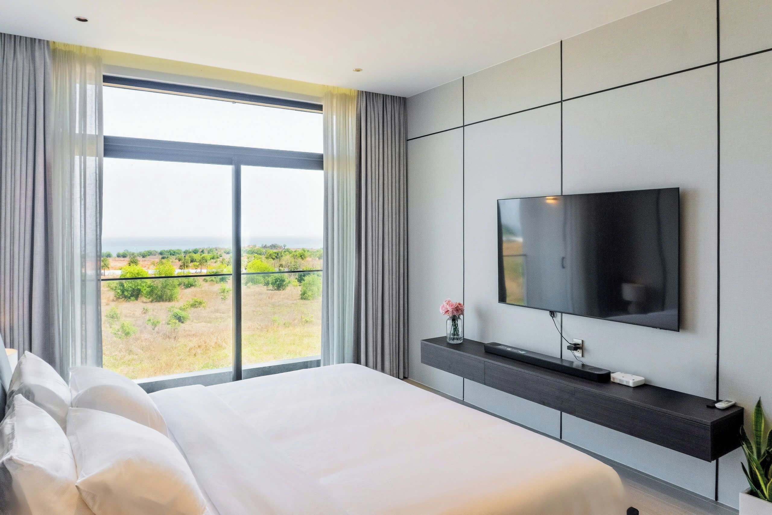 Una habitación de hotel moderna con una cama grande, un televisor de pantalla plana montado en la pared, un balcón con vistas a la vegetación y una planta pequeña en la mesita de noche.