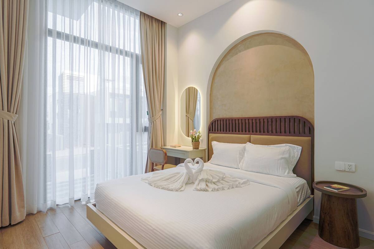 A hotel room with a large bed, a desk, a chair, a round wooden side table, and a mirror.