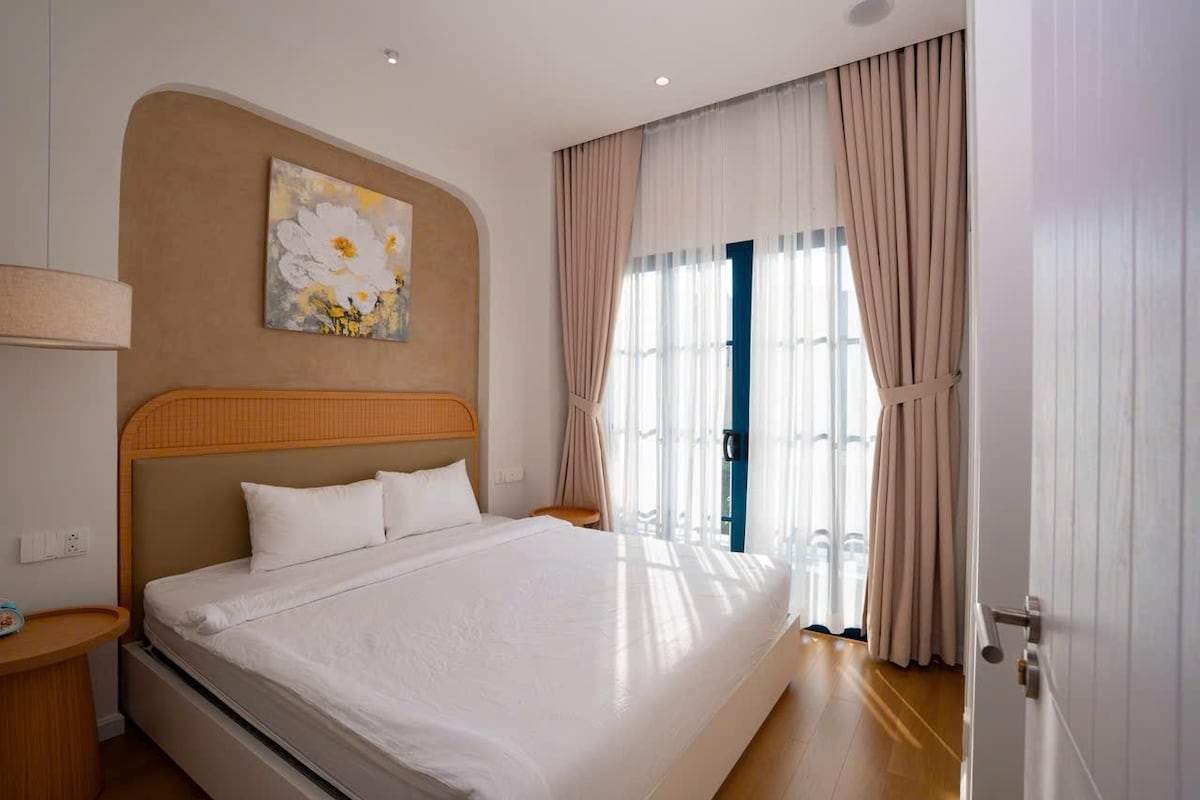 A hotel room with a large bed, a headboard, a bedside table, a wall-mounted artwork, and a window with curtains.