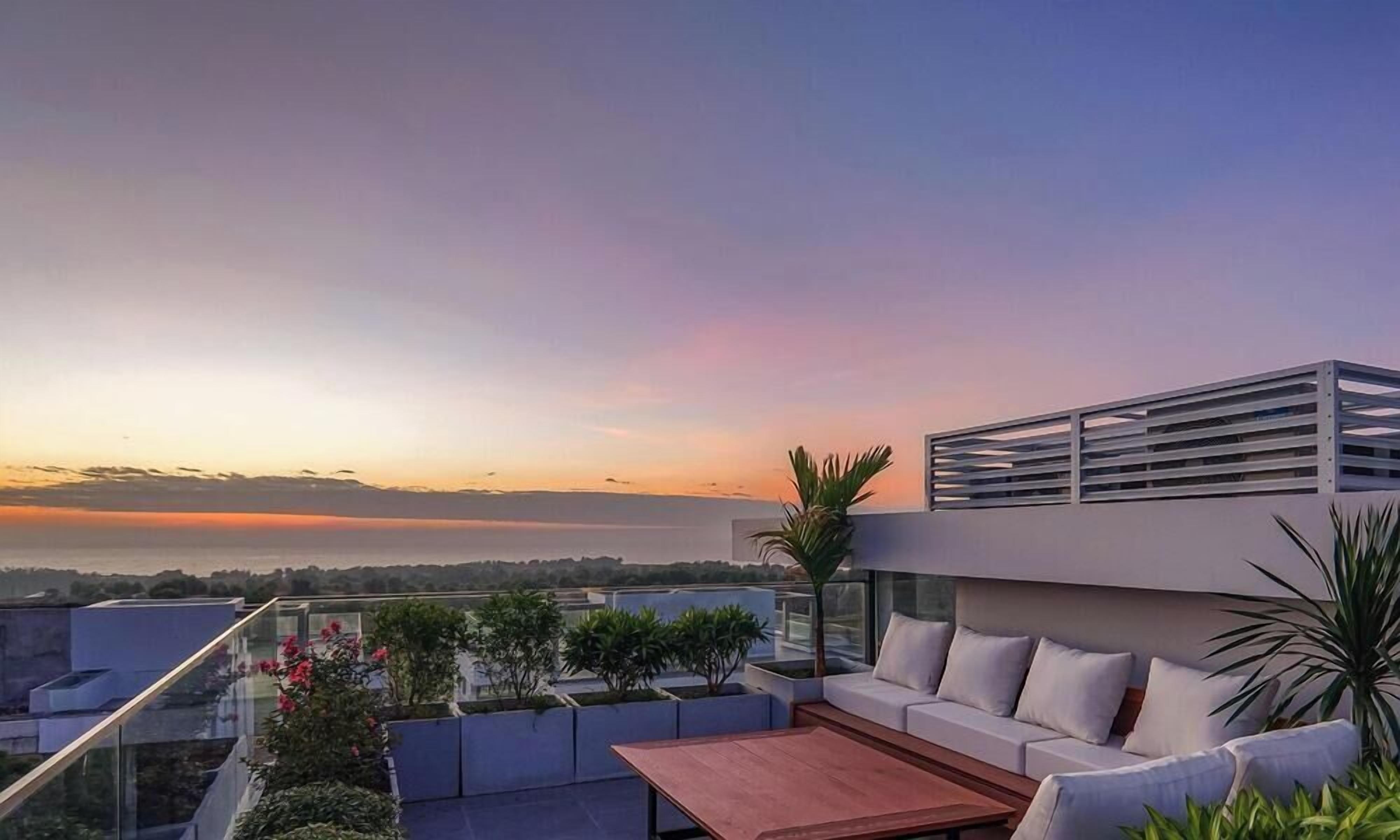 A rooftop terrace with a sofa, table, and plants, overlooking a cityscape at sunset.