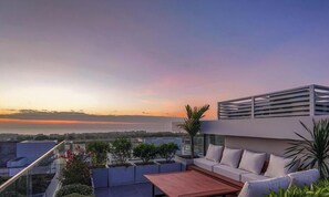 A rooftop terrace with a sofa, table, and plants, overlooking a cityscape at sunset. - Life Travel Villa - Novaworld Phan Thiet (Phan Thiet)