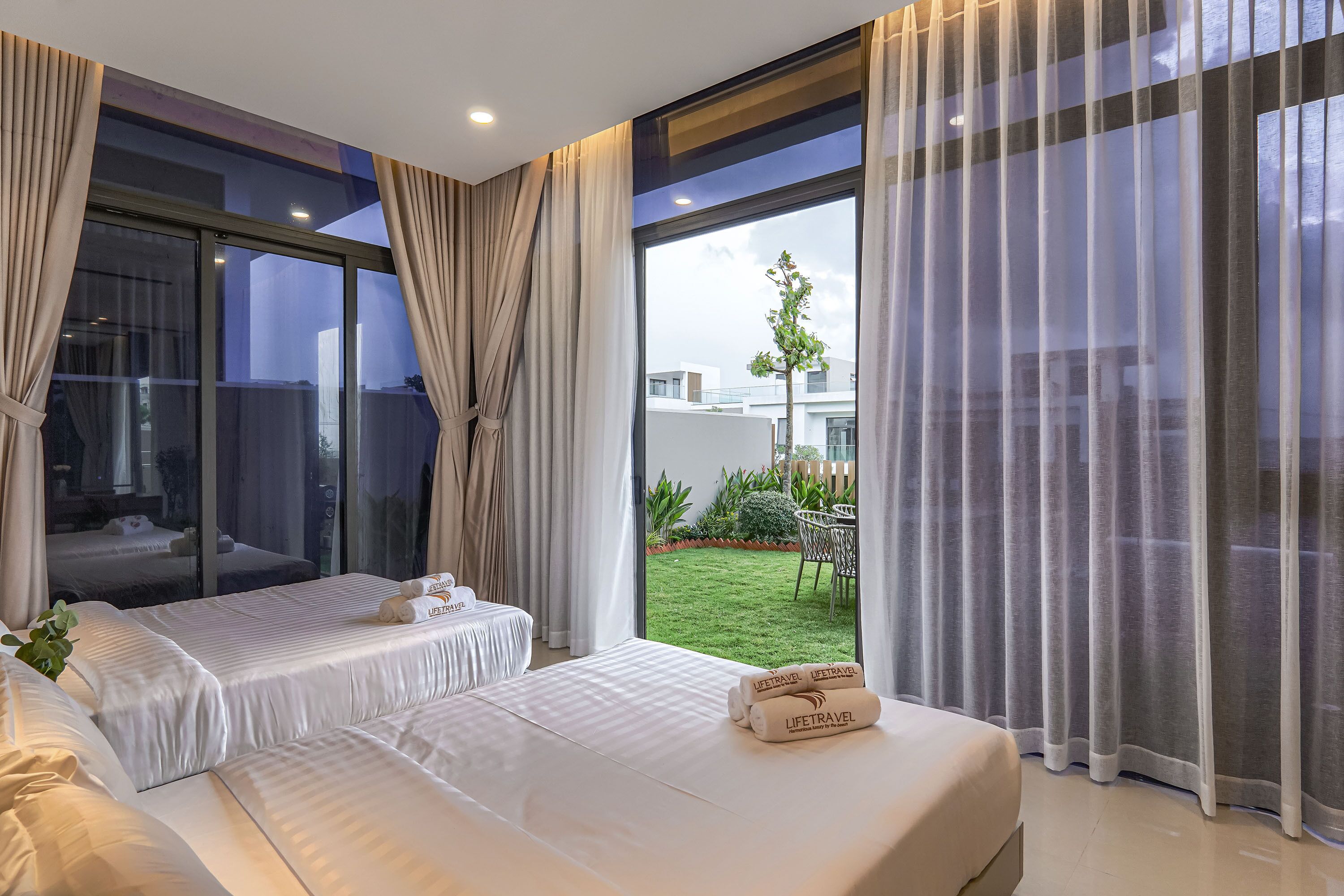 A modern hotel room with a large sliding glass door to a balcony, a bed with white linens, and a small table with a plant.