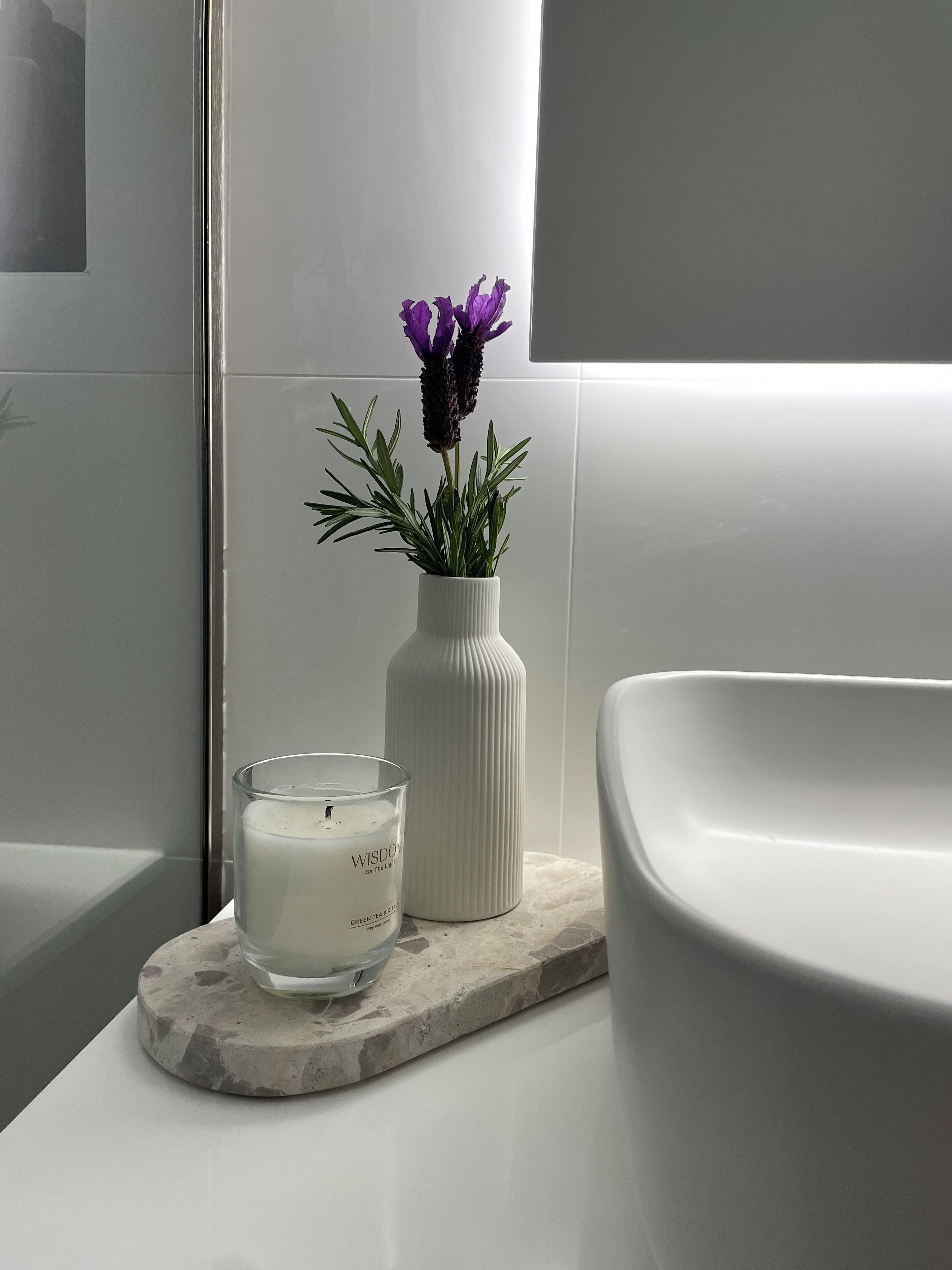 A bathroom with a white sink, a white vase with purple flowers, and a white candle on a stone slab.