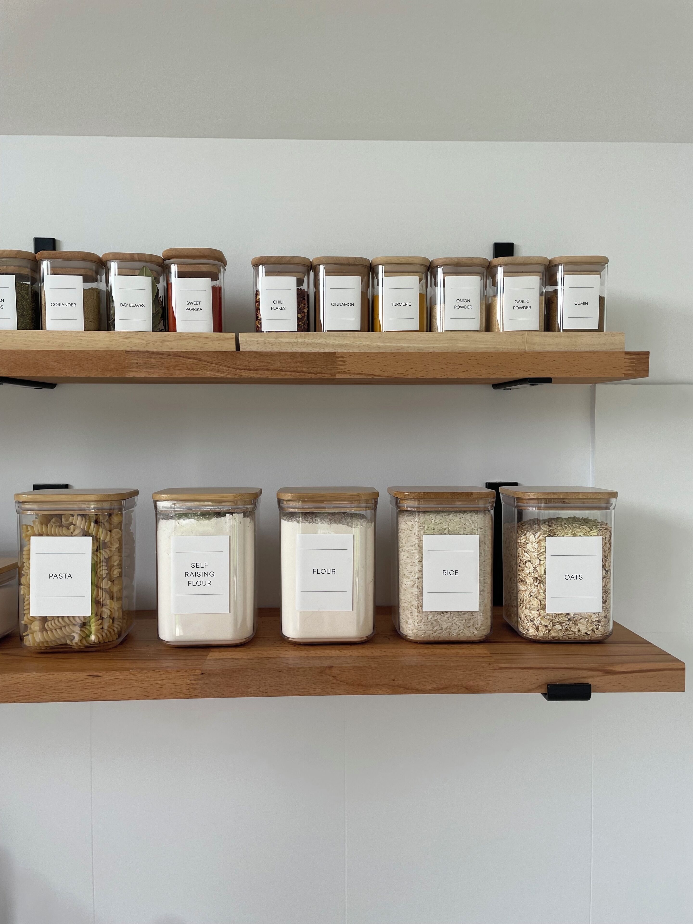 Two wooden shelves with glass jars labeled with various food items like pasta, flour, and rice.