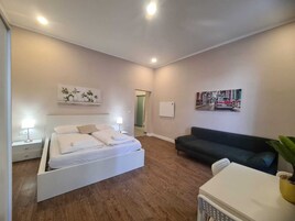 Quarto moderno com cama, mesas de cabeceira, um sofá e uma pequena mesa com uma planta.