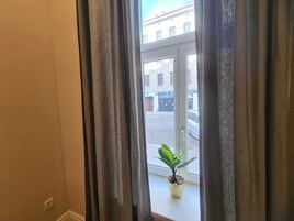 Vista pela janela, com cortinas, uma planta em vaso e um edifício com sinalização ao fundo.
