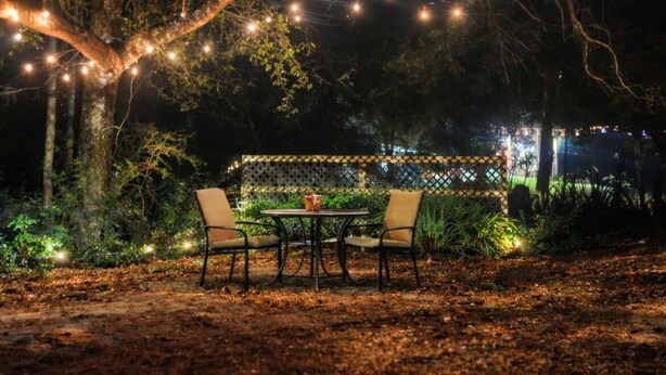 An outdoor seating area with a table and chairs under string lights, surrounded by trees.