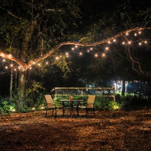 Un área de estar al aire libre con mesa y sillas bajo luces colgantes, rodeada de árboles.