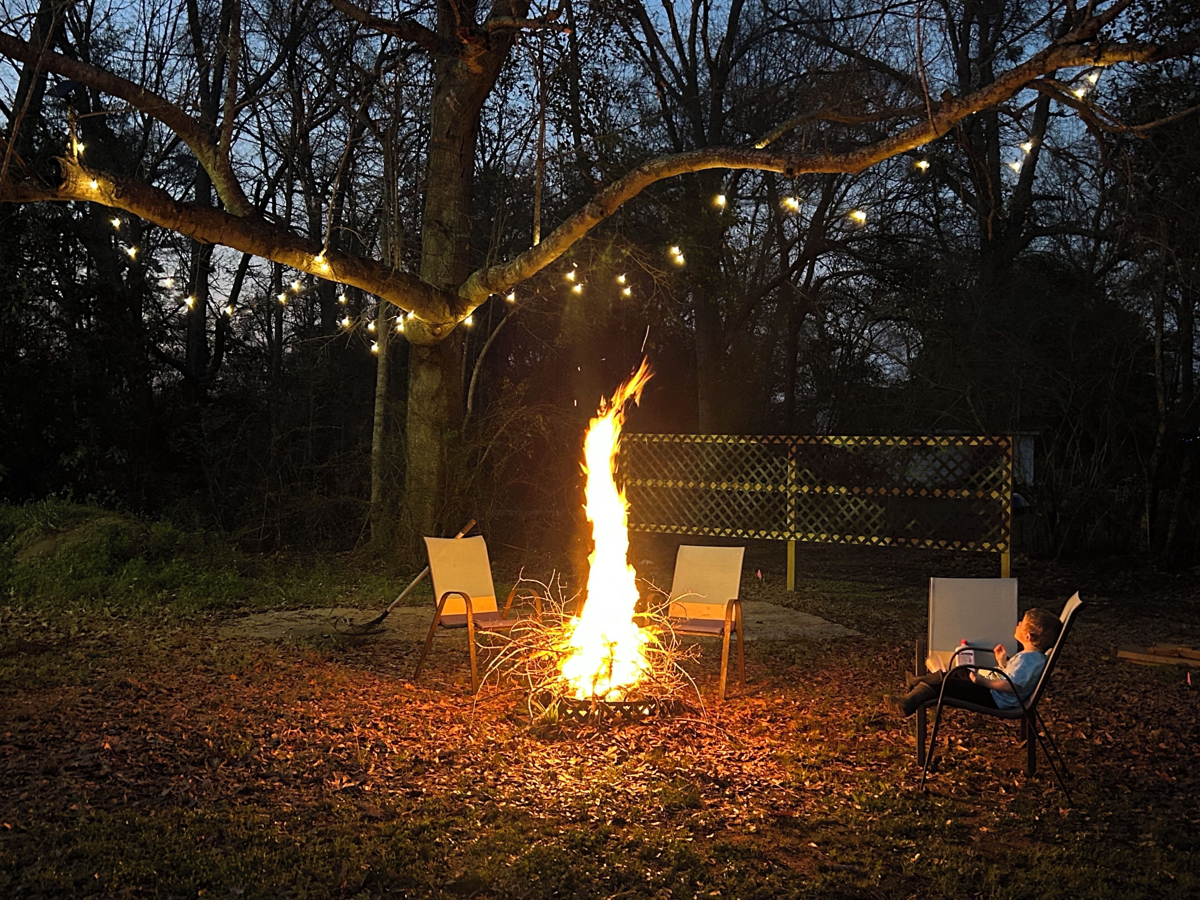 A campfire with two chairs and a table, surrounded by trees and string lights.