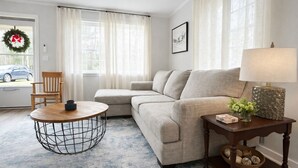 A living room with a beige sofa, a round coffee table, a wooden side table with a lamp, and a view of a car through a window.