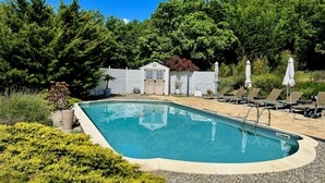 Una piscina rectangular con plataforma de madera y sillas de descanso, rodeada de vegetación y un cobertizo.
