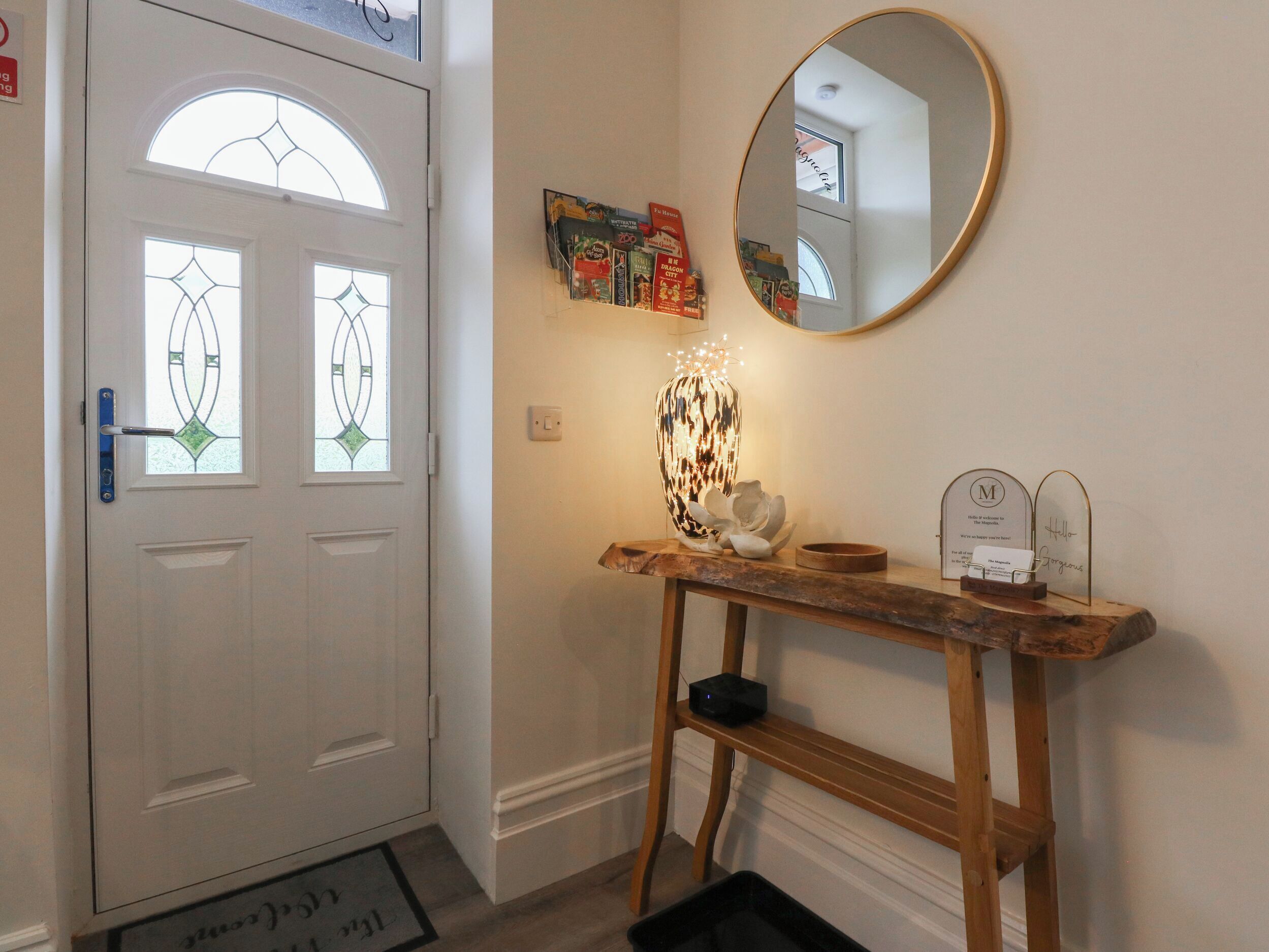 Een gang met een witte deur, een ronde spiegel, een houten console tafel met decoratieve objecten en een deurmat.