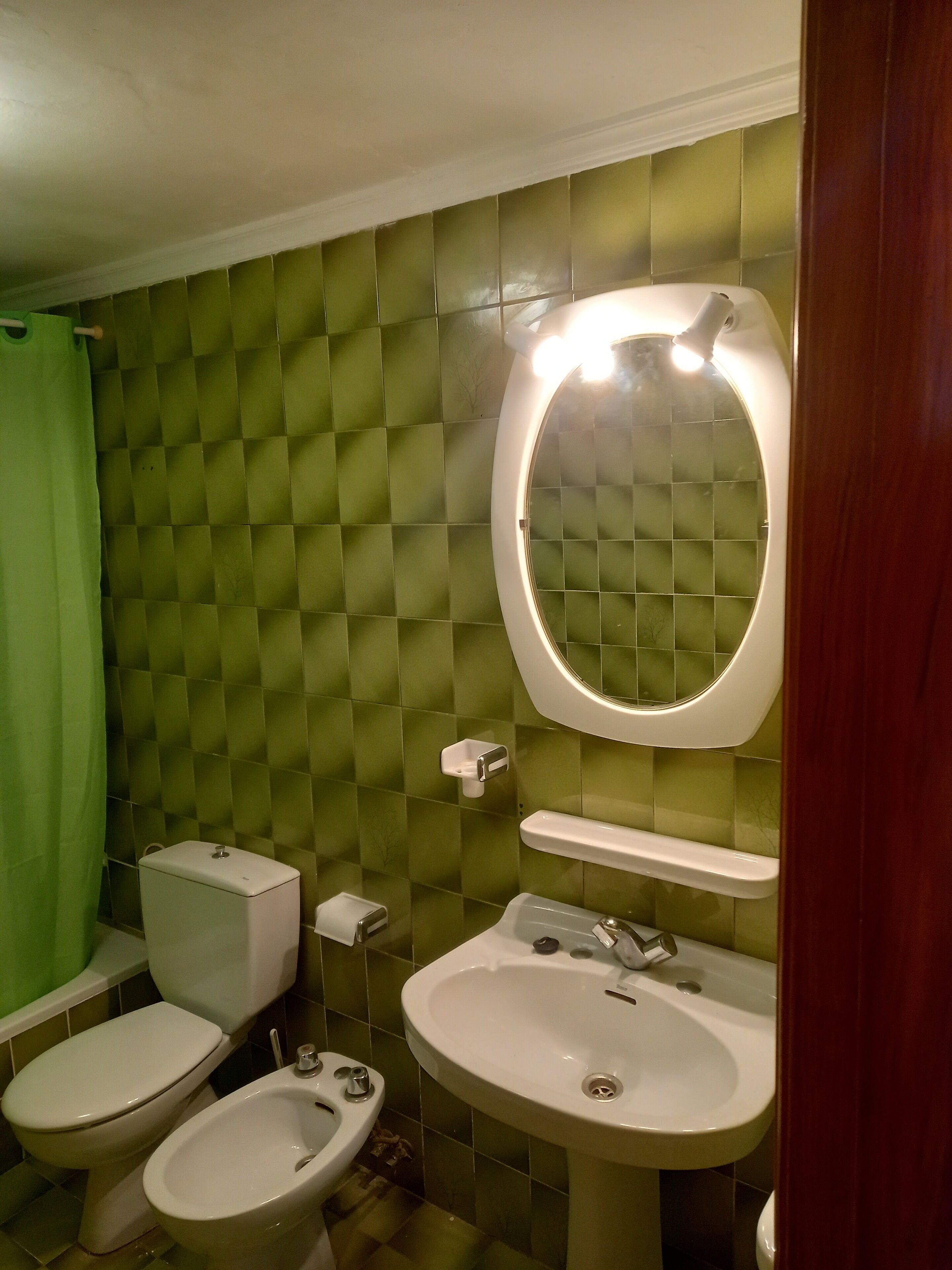 Ein Badezimmer mit Toilette, Bidet und Waschbecken, alles in Weiß, vor einer grünen Fliesenwand.