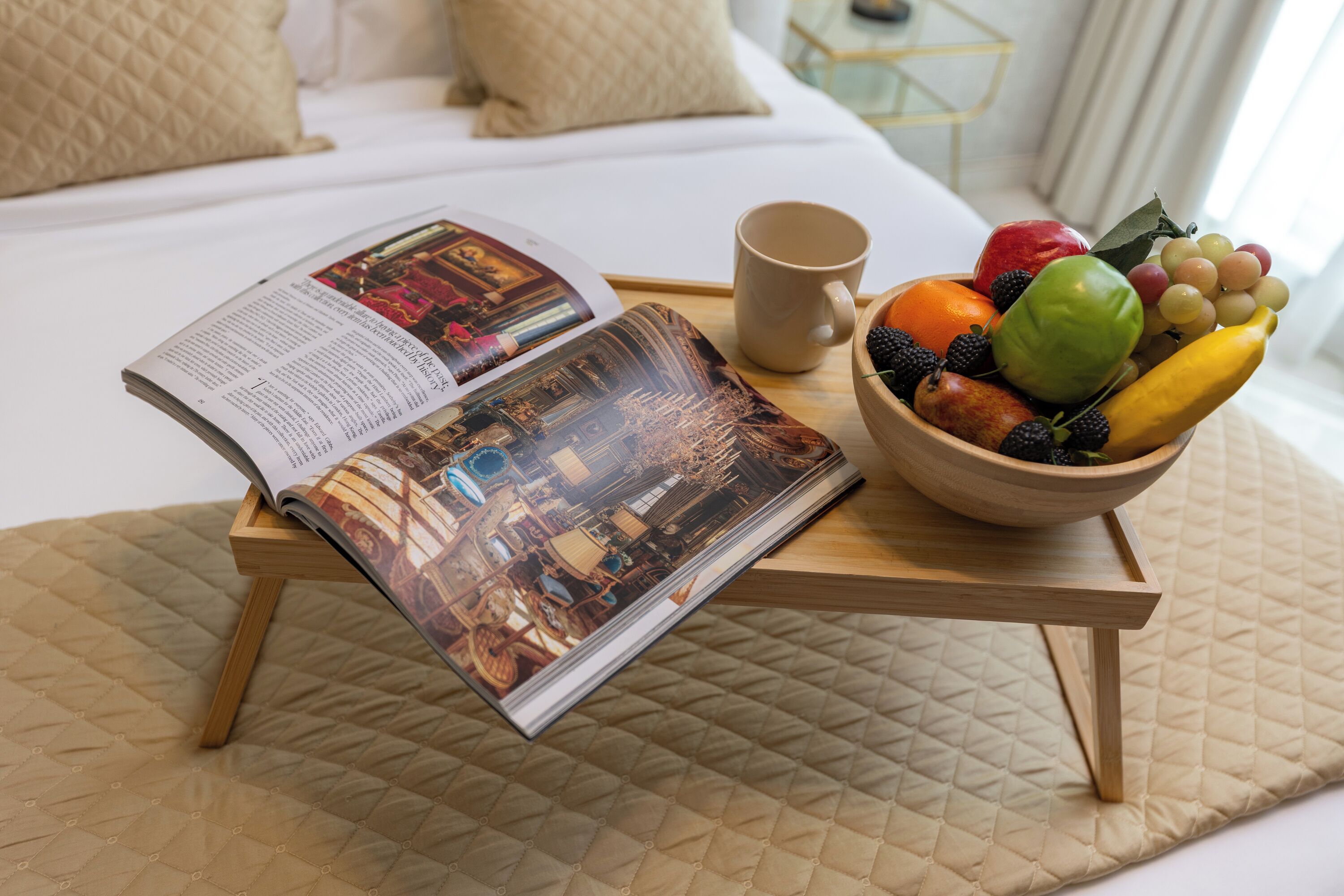 A bed with a quilted cover, a bedside table with a magazine, a bowl of fruit, and a mug.