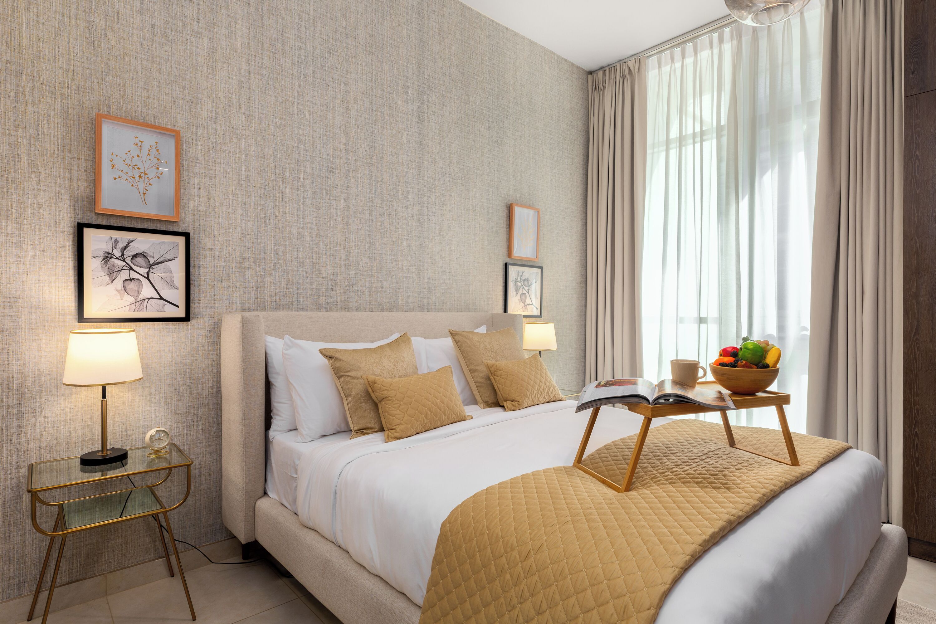A hotel room with a bed, bedside tables, a lamp, framed artwork, a bowl of fruit, and a window with curtains.