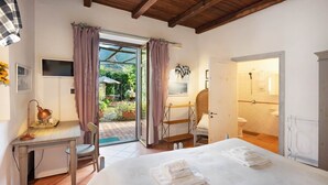 A bedroom with a bed, a desk with a lamp, a chair, a bathroom with a shower, and a view of a garden through an open door. - BARONE ANTONIO NEGRI FARMHOUSE (Fisciano)