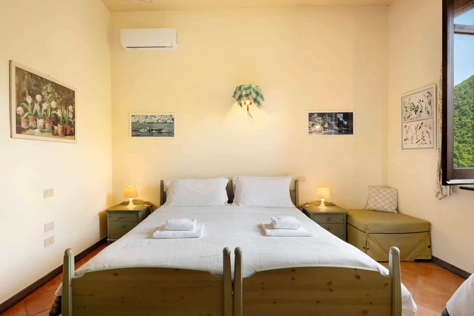 Un dormitorio con una cama doble, dos mesitas de noche con lámparas, una mesita de noche más pequeña con lámpara, una ventana con vistas a zonas verdes y una obra de arte colgada en la pared.