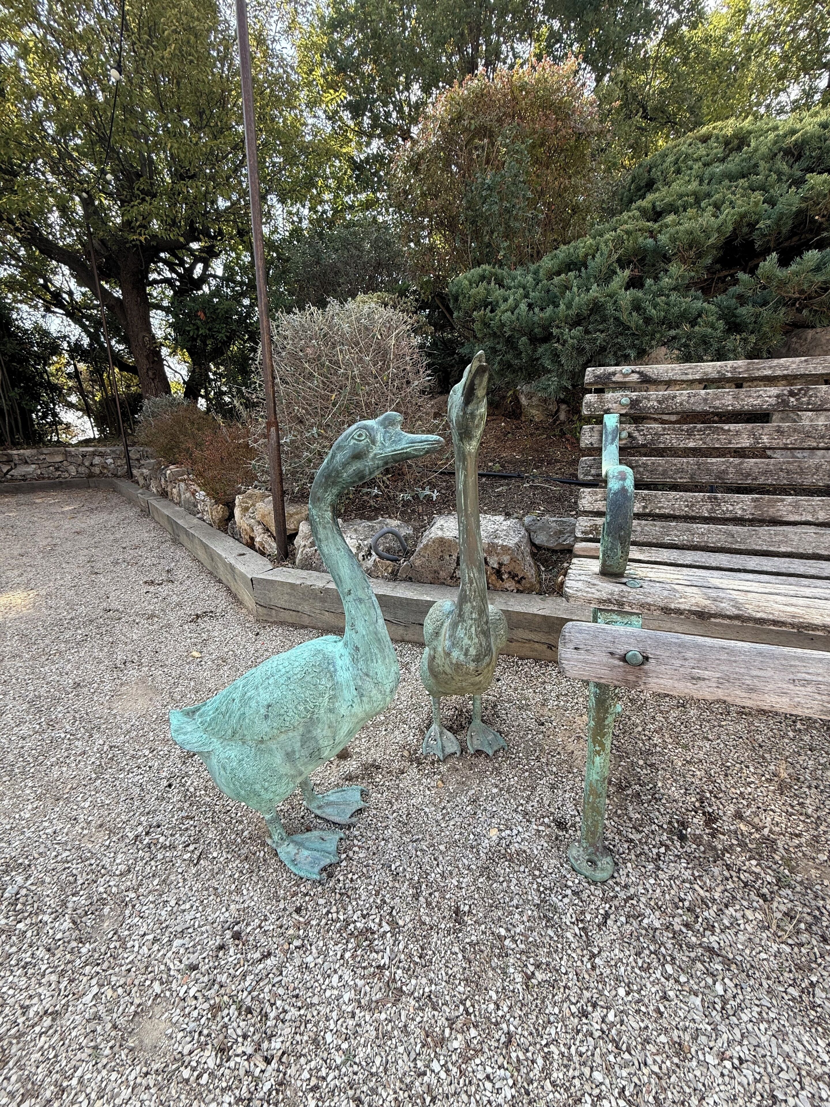 Une statue représentant deux oiseaux, dans un jardin agrémenté d’un banc en bois et d’un mur en pierre.