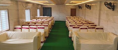 Une salle de conférence avec des rangées de chaises recouvertes de housses, une moquette verte et des ventilateurs de plafond.