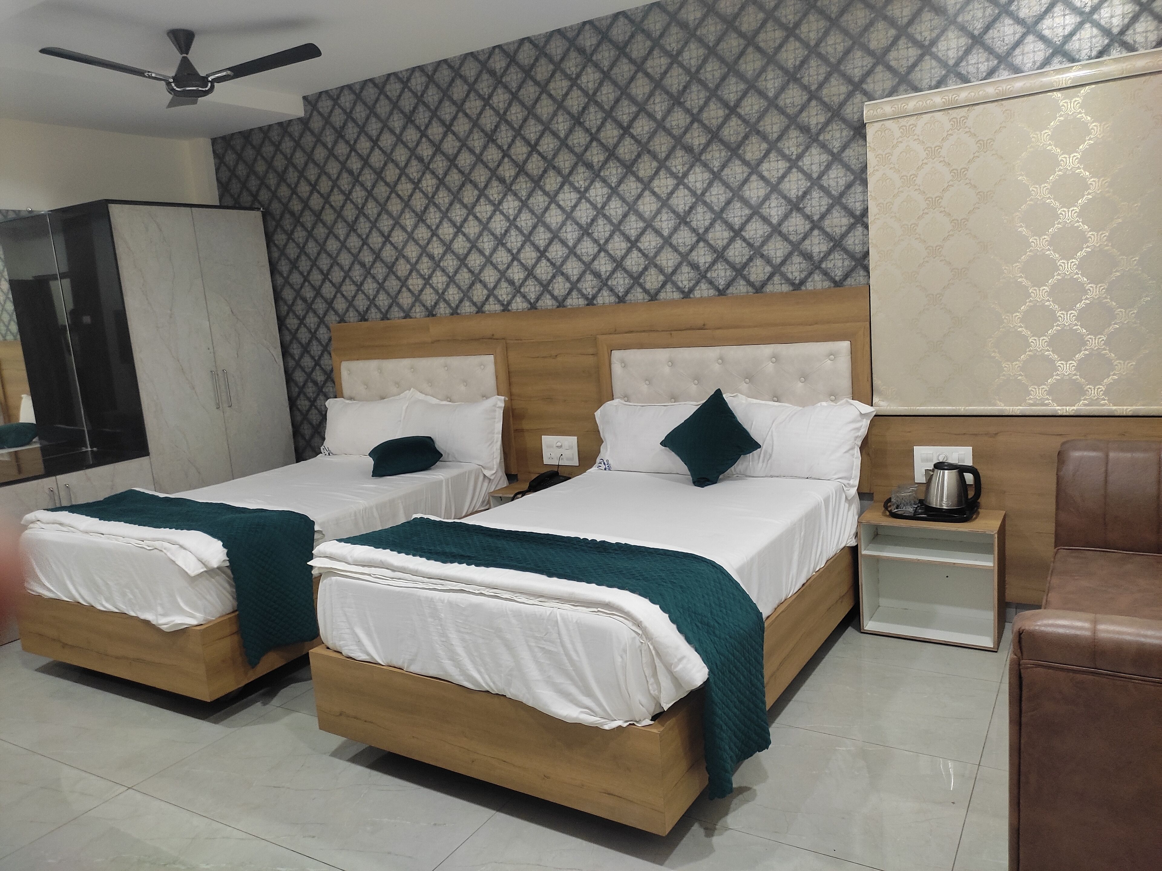 A hotel room with two single beds, a wardrobe, a nightstand with a kettle, and a ceiling fan.