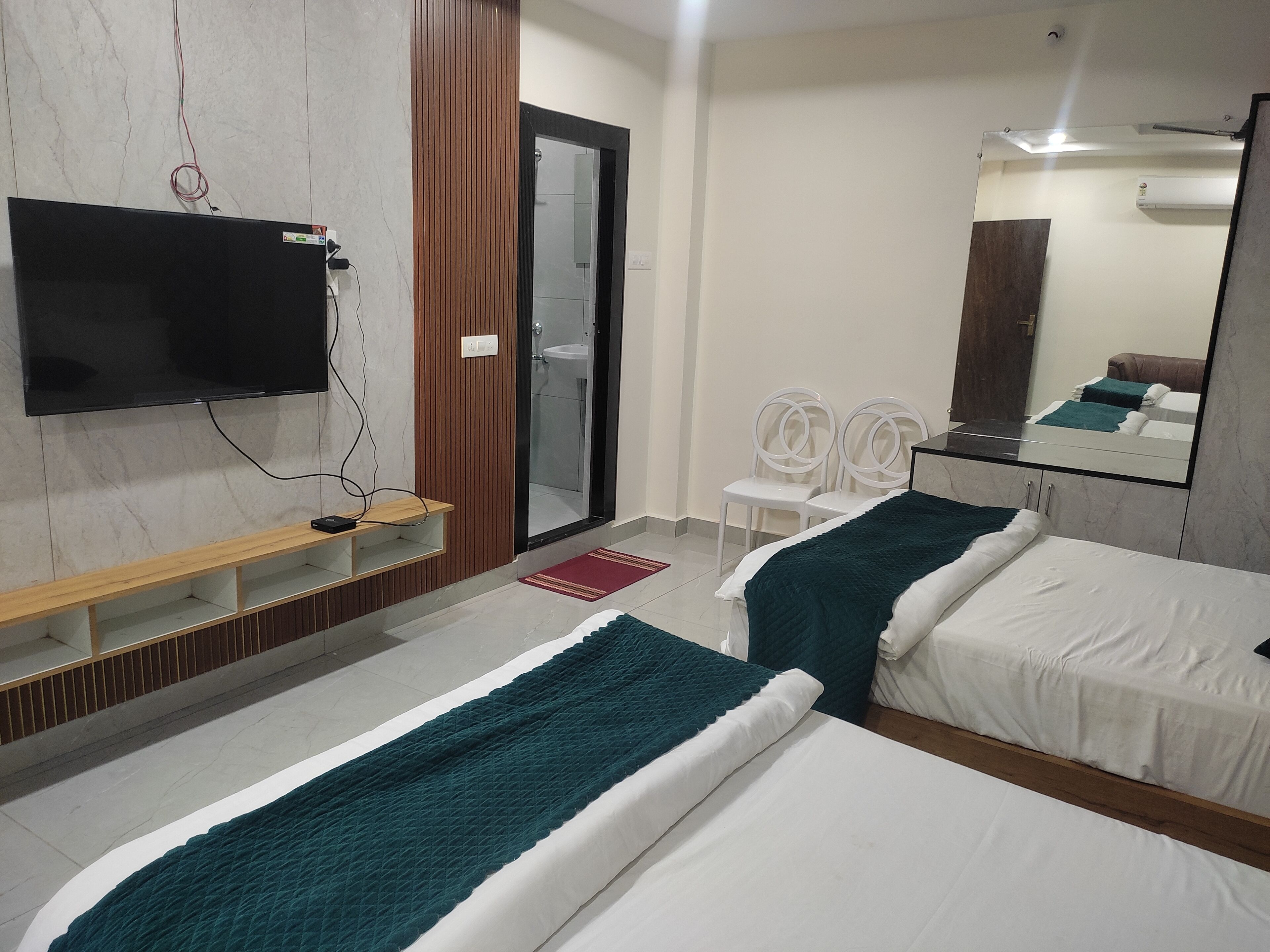 A hotel room with two beds, a TV mounted on the wall, a wooden shelf, and a bathroom visible through a glass door.