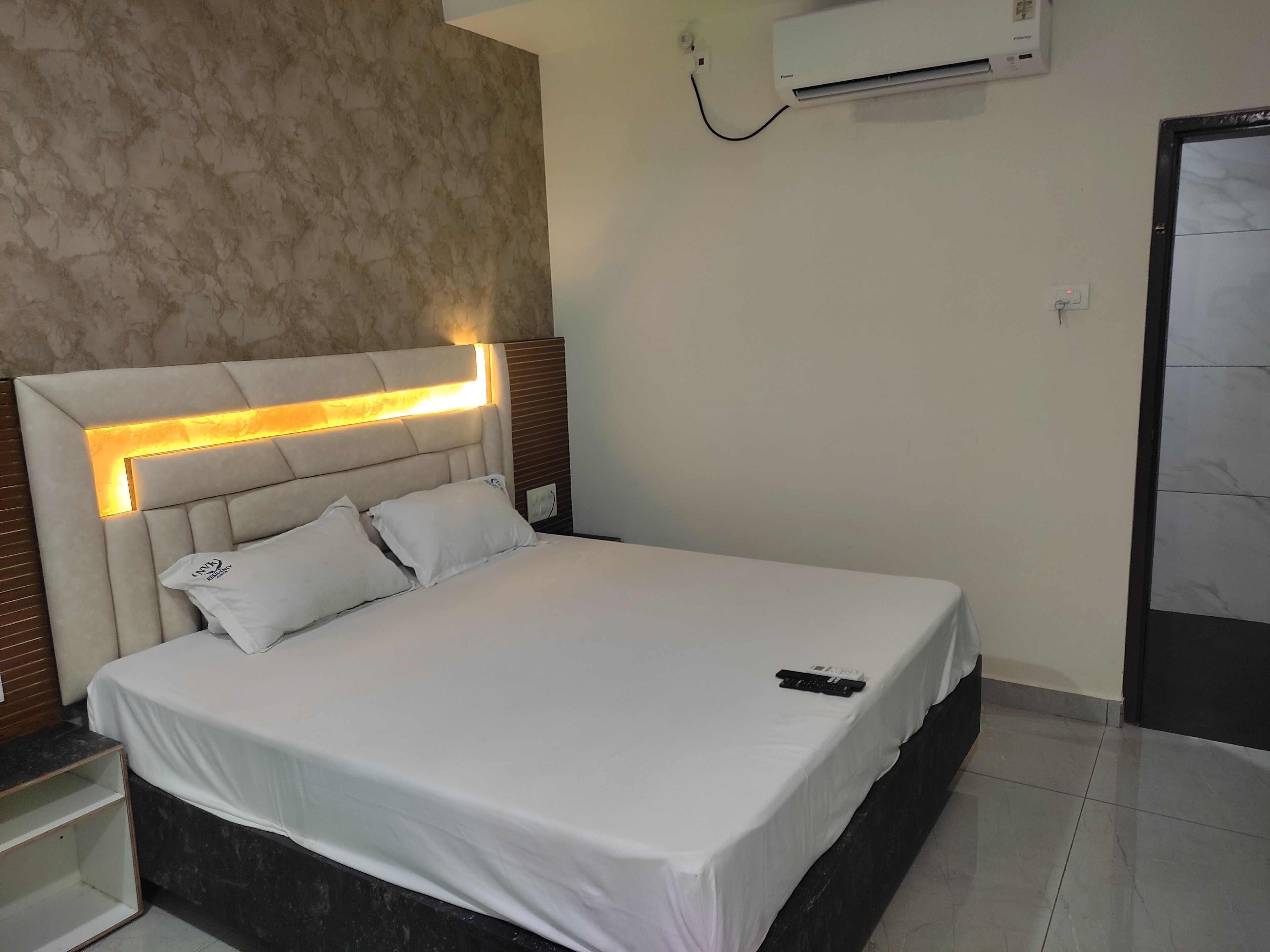 A neatly made bed with a white bedspread and two pillows, a headboard with integrated lighting, a wall-mounted air conditioner, and a door with a marble-like finish.