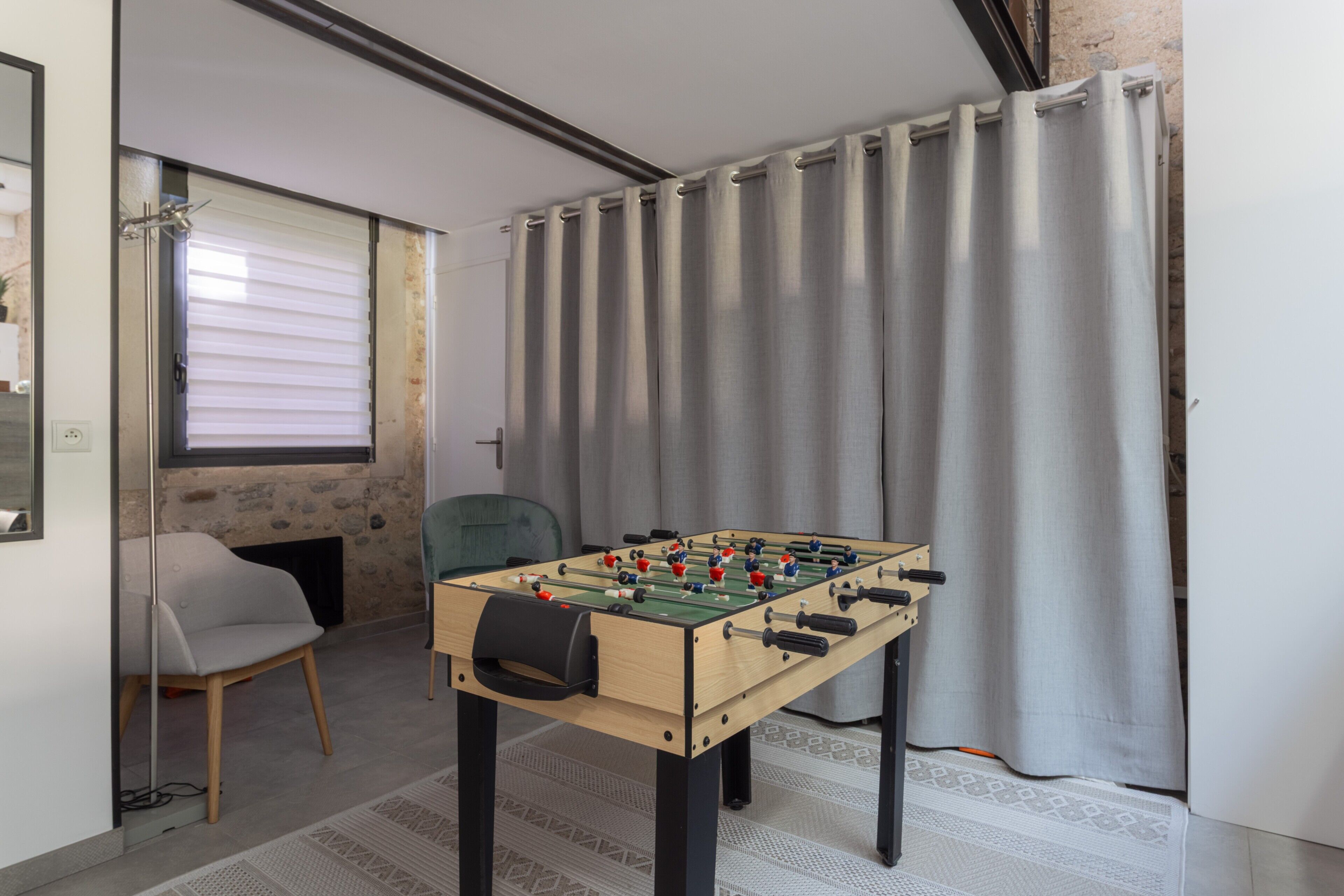 A room with a foosball table, a chair, a window with blinds, and a curtain.