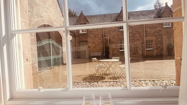 A window with a view of a courtyard, a brick building, and a table with two bottles. -  Coachman’s Cottage at Brylach Steading, Speyside (Aberlour)