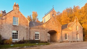 A stone building with a central archway, multiple windows, and a small tower with a clock. -  Coachman’s Cottage at Brylach Steading, Speyside (Aberlour)